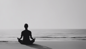 A person practices meditation, sitting cross-legged on a beach and facing the calm ocean, immersed in a peaceful and serene setting under a clear sky.