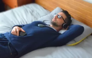 A man wearing glasses and headphones lies on a bed with one arm behind his head, holding a smartphone on his chest, appearing relaxed and possibly using the Sensate app to enhance his music experience.