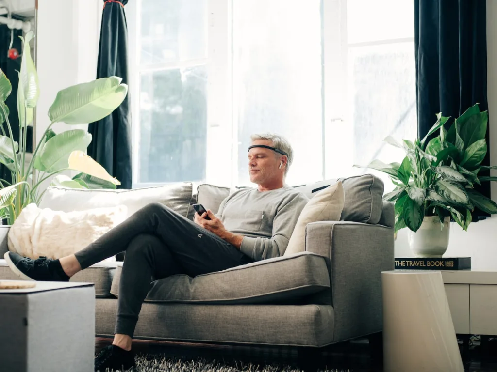 A man in casual clothes sits on a gray sofa in a bright room, wearing the Muse Your Personal Meditation Coach headband and holding a phone. Large green plants and The Travel Book are nearby as sunlight streams through the windows.
