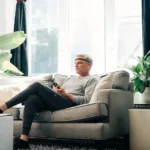 A man in casual clothes sits on a gray sofa in a bright room, wearing the Muse Your Personal Meditation Coach headband and holding a phone. Large green plants and The Travel Book are nearby as sunlight streams through the windows.