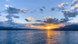 The sun sets behind mountains, casting a golden reflection on a calm blue lake. Scattered clouds dot the sky, creating a serene landscape that inspires faith and nurtures health amidst tranquil beauty. Faith and Health