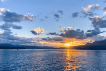 The sun sets behind mountains, casting a golden reflection on a calm blue lake. Scattered clouds dot the sky, creating a serene landscape that inspires faith and nurtures health amidst tranquil beauty. Faith and Health