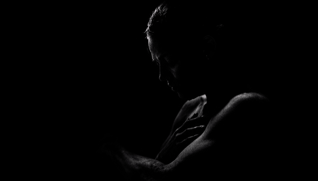 A person is shown in a dark, low-key portrait with dramatic lighting highlighting their face, shoulder, and arm. Their head is slightly bowed and one hand rests on the opposite shoulder, evoking a mood of somber introspection and quiet recovery.
