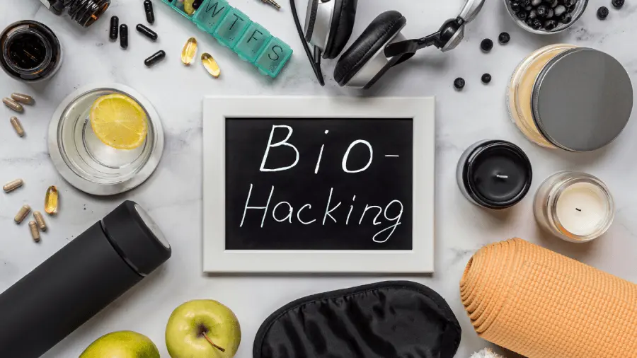 A chalkboard with Bio-Hacking written on it is surrounded by supplements, a water bottle, green apples, an eye mask, a candle, pills, a yoga mat, and headphones essentials for biohacking your physical and mental health.