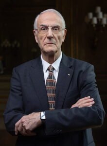 Dr. Manuel Sans Segarra, an older man in a dark pinstripe suit, patterned tie, and glasses, stands with arms crossed, looking serious. The background is dimly lit with ornate decor and a chandelier.