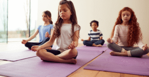 Four children sit cross-legged on purple yoga mats indoors, eyes closed and hands on their knees, calmly practicing mindfulness for children together in a bright room.